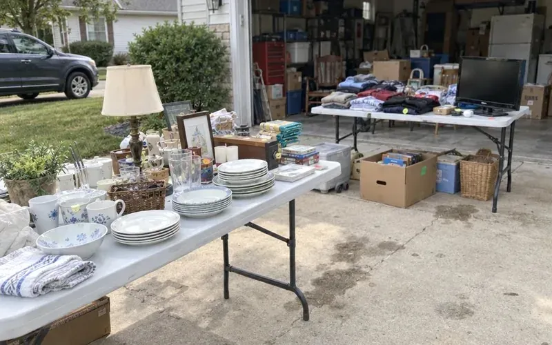 Folding tables of household items and boxes set out in a driveway for a sale before downsizing