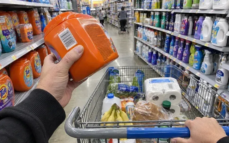 Hand holding a laundry detergent box with its barcode showing above a shopping cart