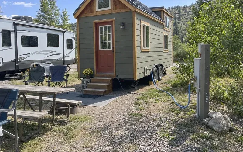 Tiny house on wheels parked at a gravel RV park site beside a utility pedestal