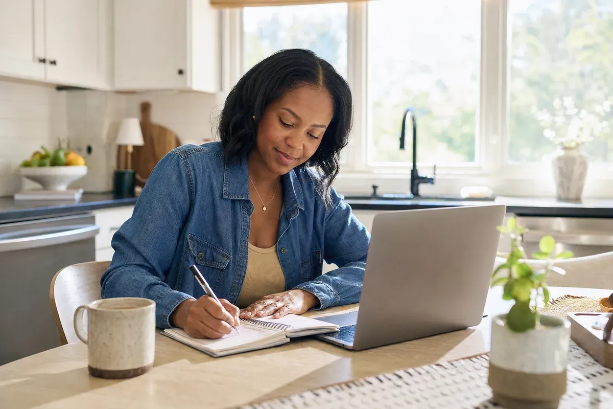 Woman at a kitchen table with a laptop and an open planner, writing down side hustle ideas over a cup of coffee