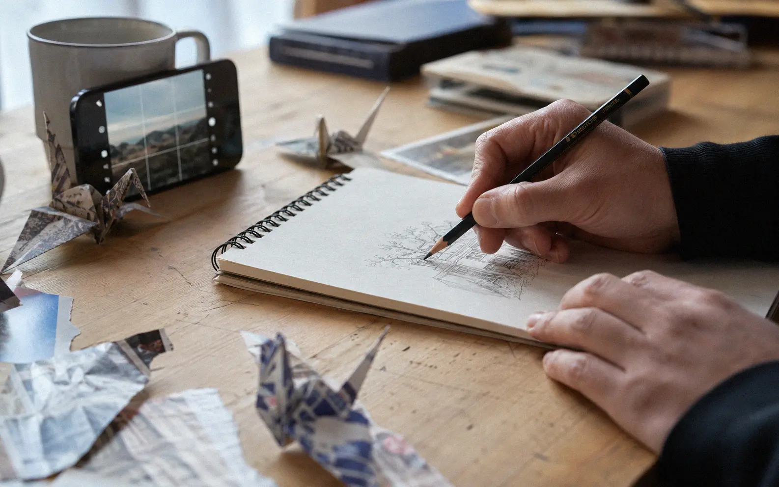 Hands sketching in a notebook surrounded by origami cranes and a phone showing photography tips